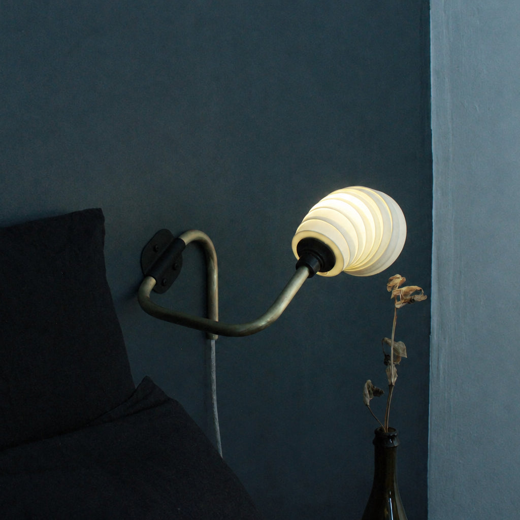 A modern wall-mounted lamp with a ribbed white globe shade and a curved dark metal arm, illuminated in a bedroom setting against a dark blue wall.