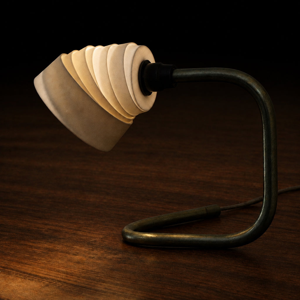 A modern table lamp with a sculptural, layered cream-colored shade and a dark, curved brass base, shown illuminated on a dark wood surface.